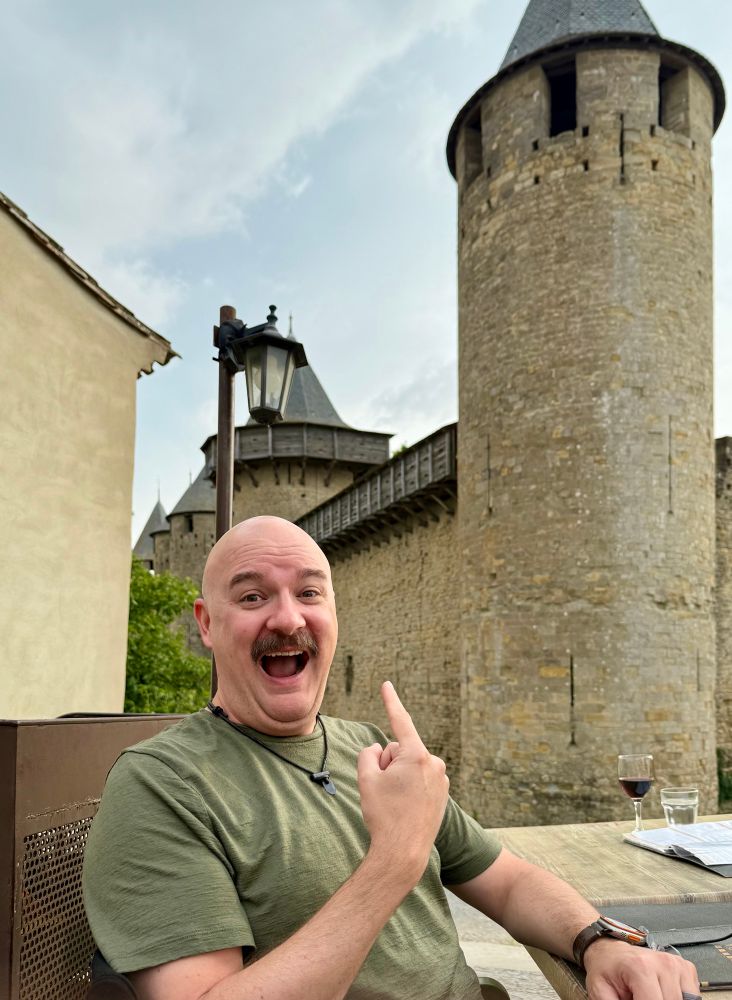 A rather giddy Michael Livingston points excitedly at the fortifications of Carcassonne. He has a requisite mustache. 