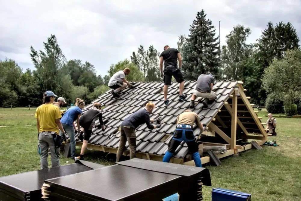 Mehrere Menschen arbeiten gemeinsam auf einem Übungsdach im Freien, um Solarpanels zu installieren. Sie tragen Arbeitskleidung und sichern sich auf dem Dach, während andere am Boden mithelfen. Die Umgebung ist grün und von Bäumen umgeben – eine praxisnahe Lernsituation im Rahmen eines Solar-Workshops.