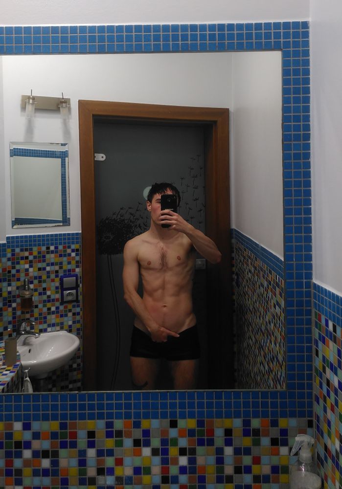 white trans man in swimshorts takes selfie in a bathroom mirror, his face is mostly covered by his phone. He is grabbing his crotch, hand inside his pants. 