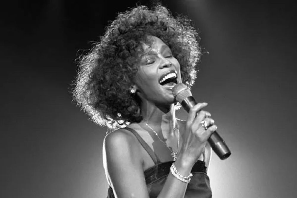 B&W photo of Whitney Houston, singing while holding a microphone