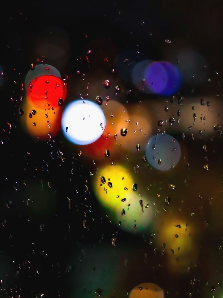 Raindrops on a window are backlit by the distant lights from the street below. 