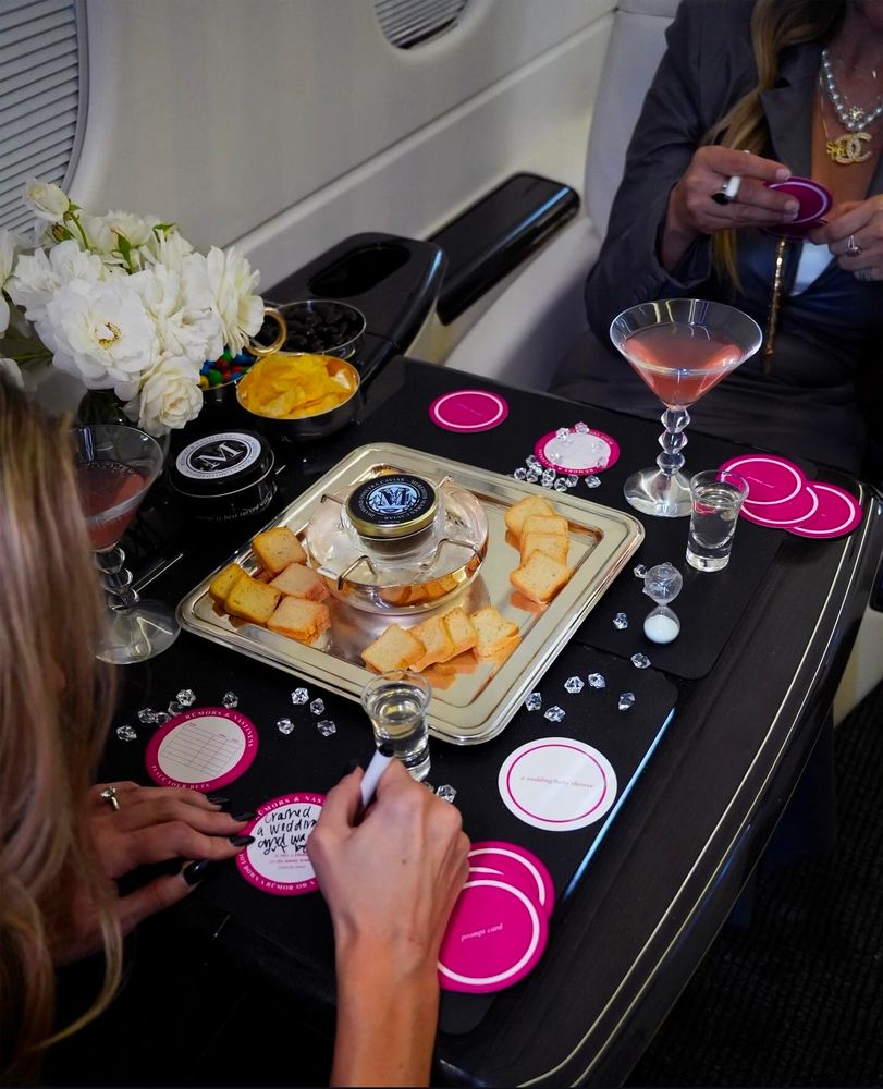 Two ladies play the Meredith Marks board game Rumors and Nastiness. On a table, a caviar tin sits surrounded by croutons. There are shot glasses, martini glasses, and fake diamonds on the table as well.
