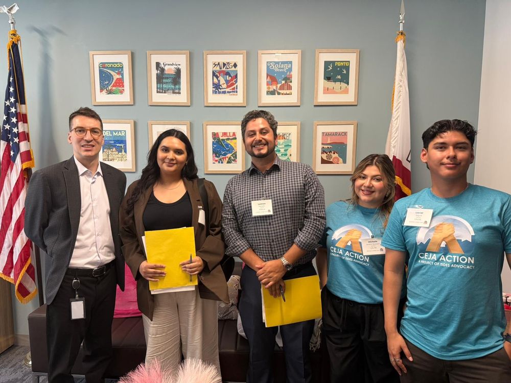 Five people standing inside the Capitol building after a lobbying meeting to discuss environmental justice priorities. Two are wearing blue CEJA Action t-shirts. Behind them are a US and California flag, and framed photos hang from the wall.
