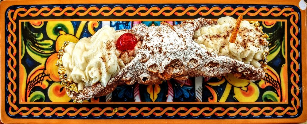 Auf einem folkloristisch orangefarbenen Tablett mit kitschigem Dekor thront ein mit Sahne, Puderzucker und einer einsamen Kirsche verziertes Gebäck, das vermutlich eine Cannolo sein möchte. Das Ensemble wirkt wie eine barocke Ode an die Farbe Orange, bei der selbst die italienische Kachel im Hintergrund versucht, mit dem Dessert in puncto Opulenz zu konkurrieren.