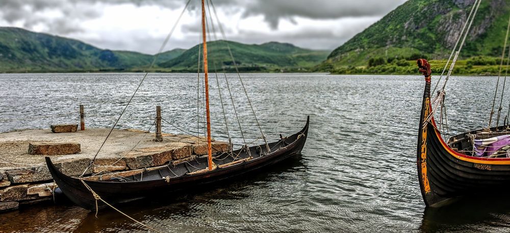 Zwei traditionelle Wikingerschiffe dümpeln malerisch am Ufer eines norwegischen Fjords auf den Lofoten, umgeben von dramatischen grünen Bergen und typisch nordischem Wolkenhimmel. Während moderne Touristen mit Kreuzfahrtschiffen und Elektroautos anreisen, harren diese historischen Fortbewegungsmittel geduldig aus – vermutlich ohne WLAN und Sitzheizung.