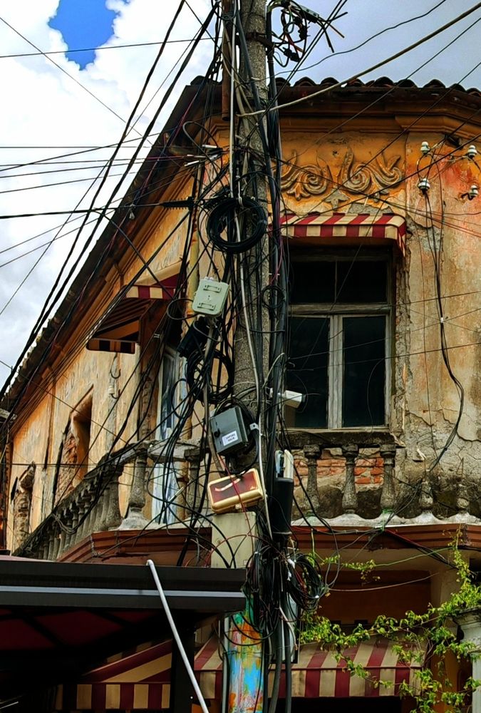 Das Bild zeigt ein altes, verwittertes Gebäude mit kunstvoller Fassade, das von einem wahren Chaos aus Stromkabeln, Telefonleitungen und verschiedenen Verteilerkästen überwuchert wird. Der Utility-Pole vor dem Haus gleicht einem technischen Spinnennetz, bei dem man sich fragt, ob hier noch jemand durchblickt oder ob das Telefon einfach per Zufall funktioniert.