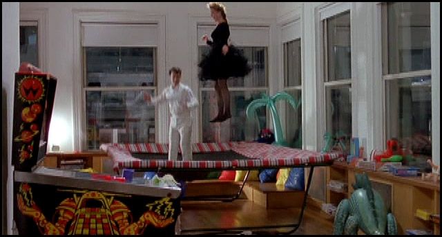 Loft mit riesigem Trampolin, auf dem 2 erwachsene Menschen gleichzeitig hüpfen, Mann in Weiß und Frau in Schwarz. Rundherum Spielzeug.