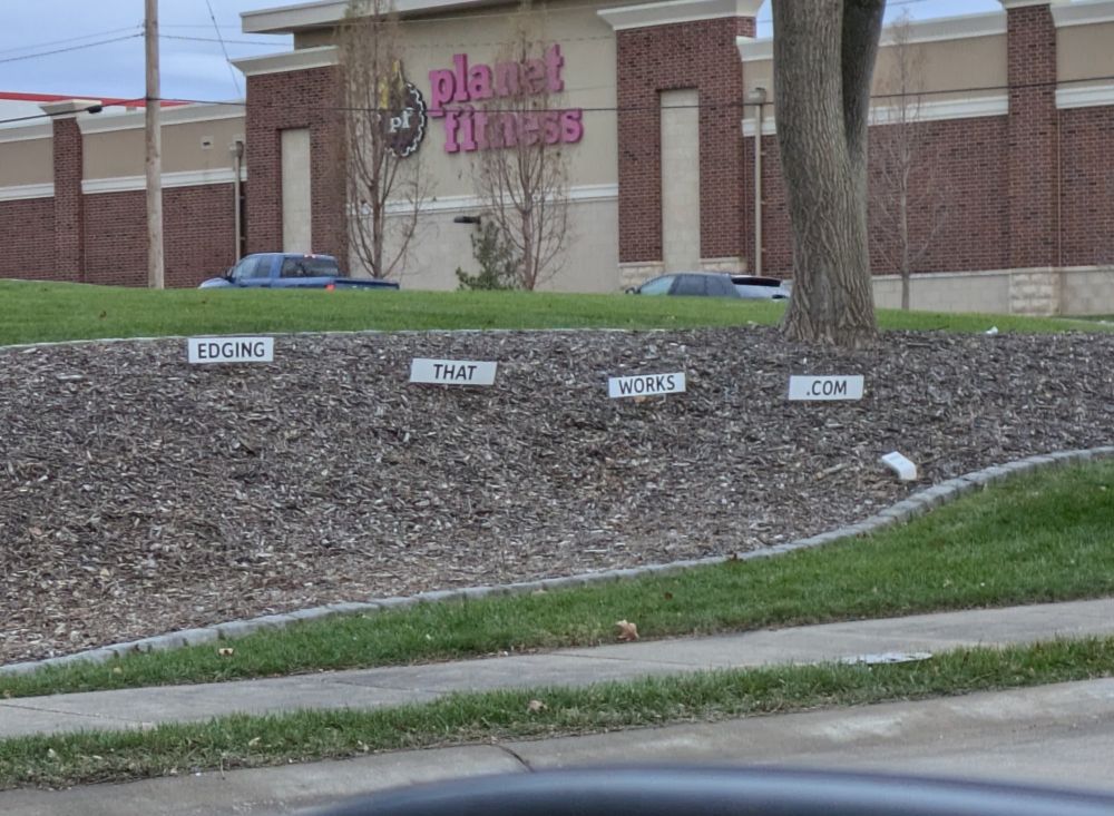 A series of four signs planted into the side of a hill on the side of the road. Across the signs, the words "Edging that works .com" can be read. Planet Fitness stands menacingly in the background. 
