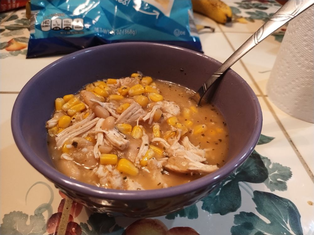 A purple bowl sits on a tiled table. Visible in the bowl is shredded chicken, corn, broth, and flecks of spice.