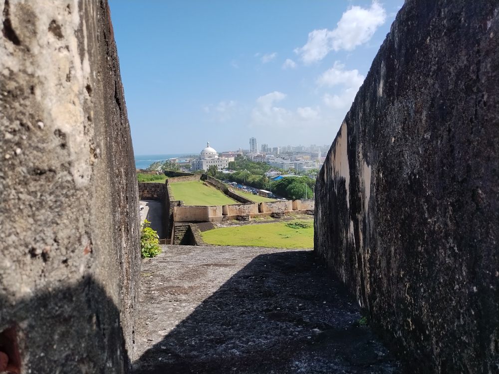 Looking between crenellations of Castillo San Cristobal, we see more of the castle below, green fields surrounded by aging stone, and San Juan in the farther background.