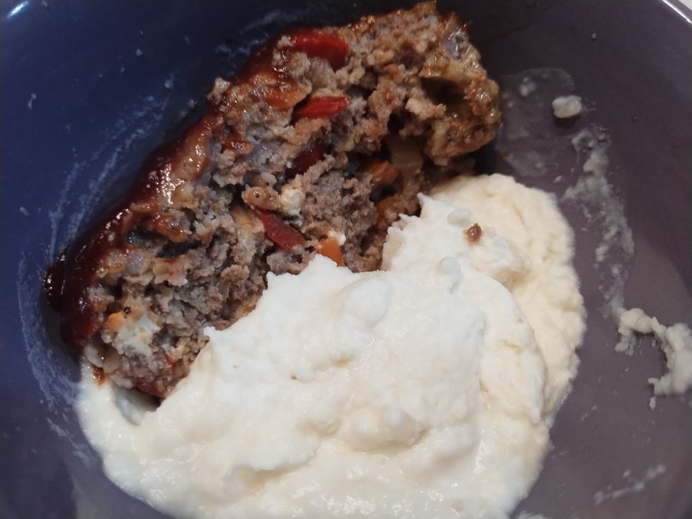 Meatloaf showing red peppers and feta, next to mashed potatoes in a purple bowl.