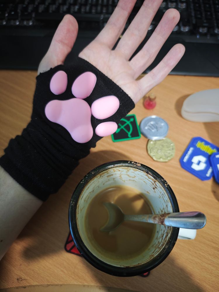 Photo of Noma's hand wearing a arm warmer that's not fully stretched over their arm, the palm featuring a pink set of paw pads for the beans part of the coffee beans. Besides the racc's hand beans, there's what's left of a cappuccino in a small mug, for the coffee part of coffee beans. A few trinkets lie on the desk, a tiny potion bottle, filled with a pinkish potion sized for a micro, a ranger token, featuring a bow and arrow in green on black, a couple of 3D printed super credits, a 3D printed NFC silver coin with a wolf, and a golden coin from fauntastic.