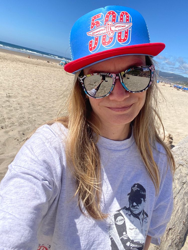 Blonde gal with a blue Indy 500 hat and a Dale Earnhardt shirt on a beach in Oregon