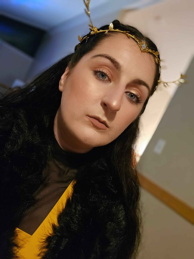 bsky user liesandarbor in a yellow and black mesh dress, fur capelet, and stag horned diadem