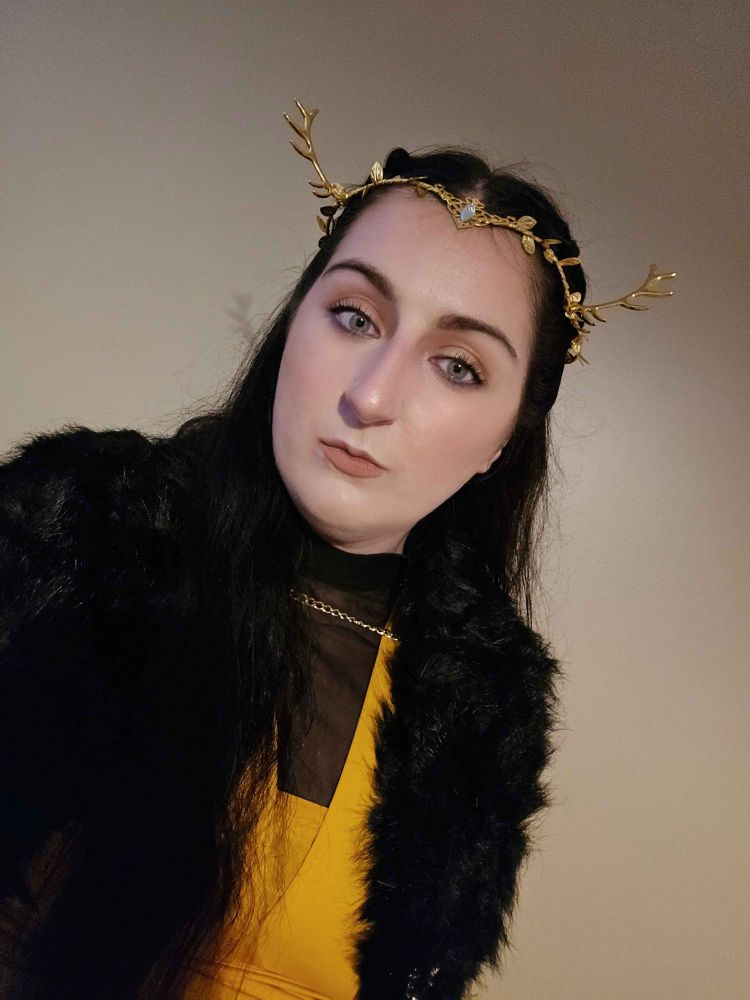 bsky user liesandarbor in a yellow and black mesh dress, fur capelet, and stag horned diadem