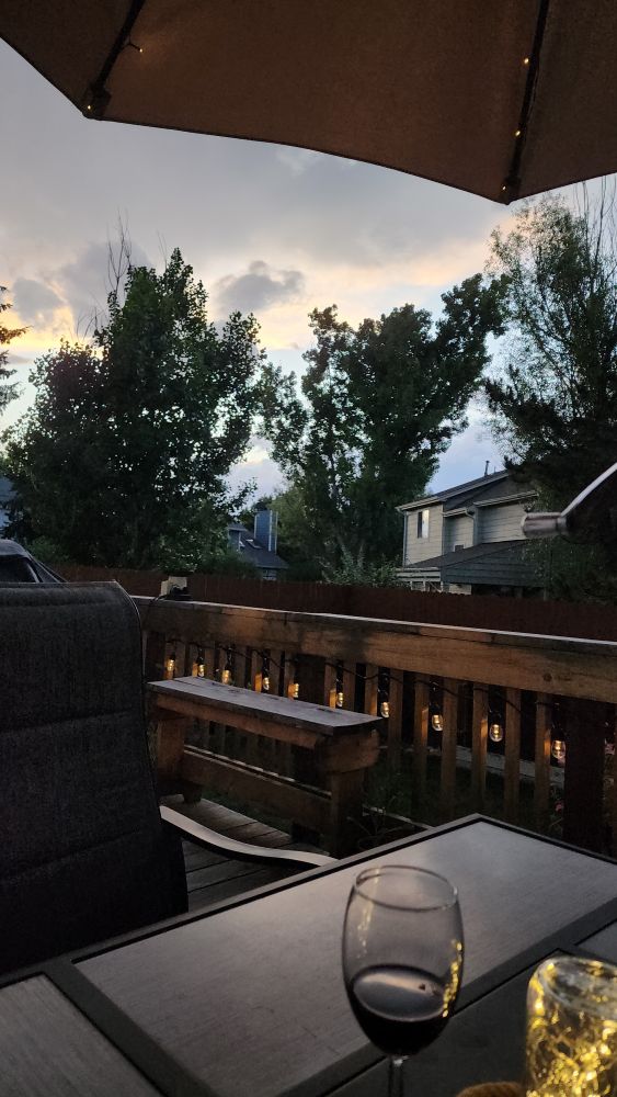 Rain clouds at sunset, a string of patio lights lines the deck rail. Under a lighted umbrella, a glass of dark burgundy colored sake in a glass sits next to fairy lights in a jar.