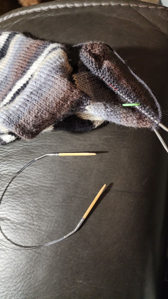 Photo of a hand knit sock in progress on a new metal needle. The old wooden one is sitting in the foreground. 