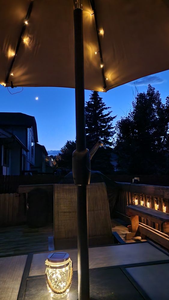 A crescent moon shines in the distance. The view from under a lighted umbrella, deck lights glowing off to the side. 