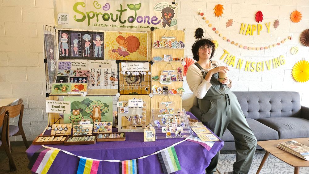 Beansprout standing next to the Sproutdone booth with a coffee in their hands.  The colorful display has art prints, stickers, button pins, and more.  The sign in the back says Happy Thanksgiving.