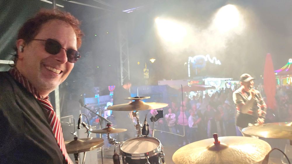 Blick von der Bühne auf das feiernde Konzertpublikum. Der lachende Drummer ist im Vordergrund.