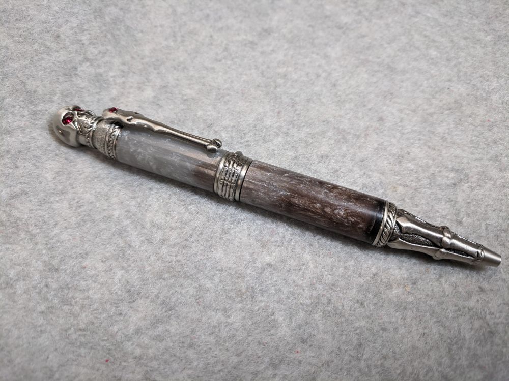 A ballpoint pen on a gray background. It has antique pewter skull and bones themed metal hardware and a resin barrel. The barrel is pale silver at the top, fading evenly to gray brown at the bottom.