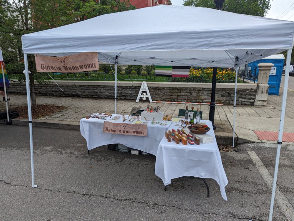 A craft show booth of two tables with white table cloths set up under a white shade canopy.