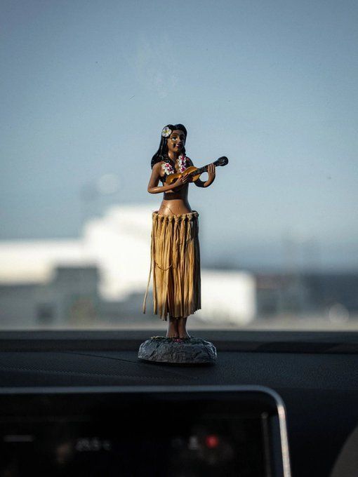 

A Hawaiian hula girl dashboard bobblehead with a grass skirt and flower lei playing a ukulele, placed on a car dashboard with a blurred outdoor background.
