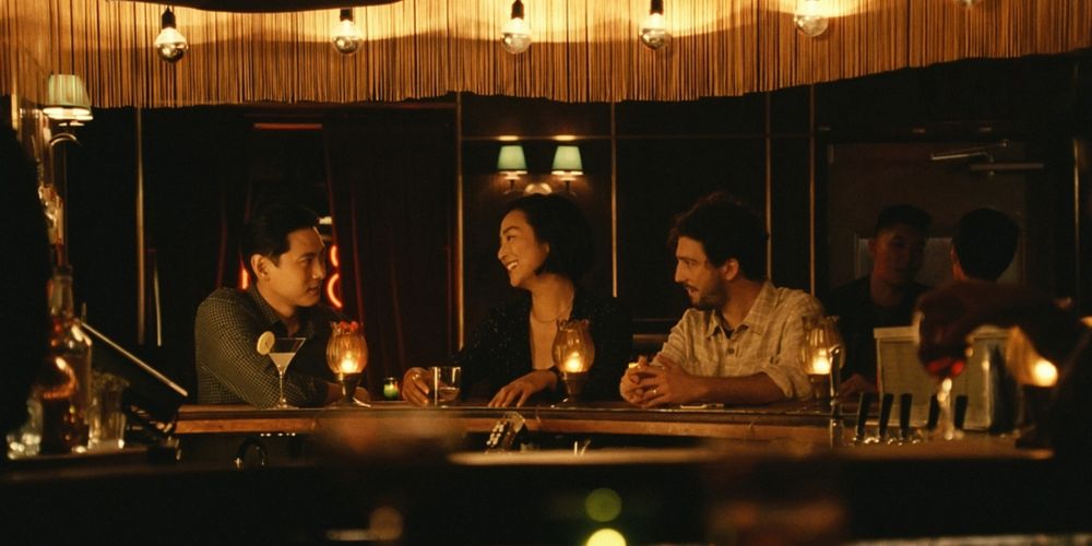 A screenshot from the movie "Past Lives". Three people (a Korean man and woman, and a Caucasian man) are sitting at a restaurant/bar and talking. 