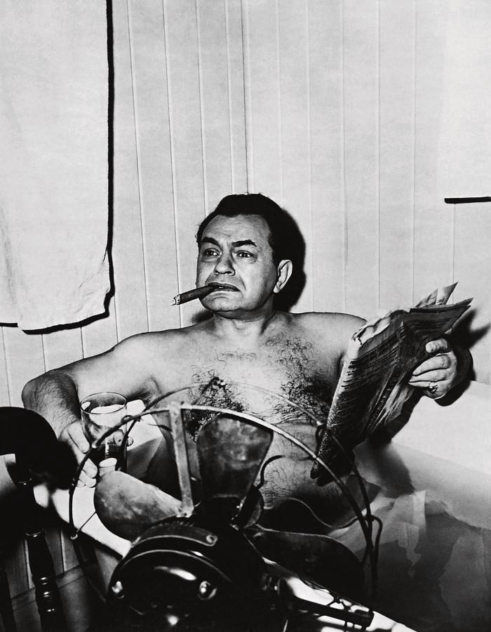 Edward G. Robinson sits in a bathtub with a cigar in his mouth, a drink in one hand, and a newspaper in the other.