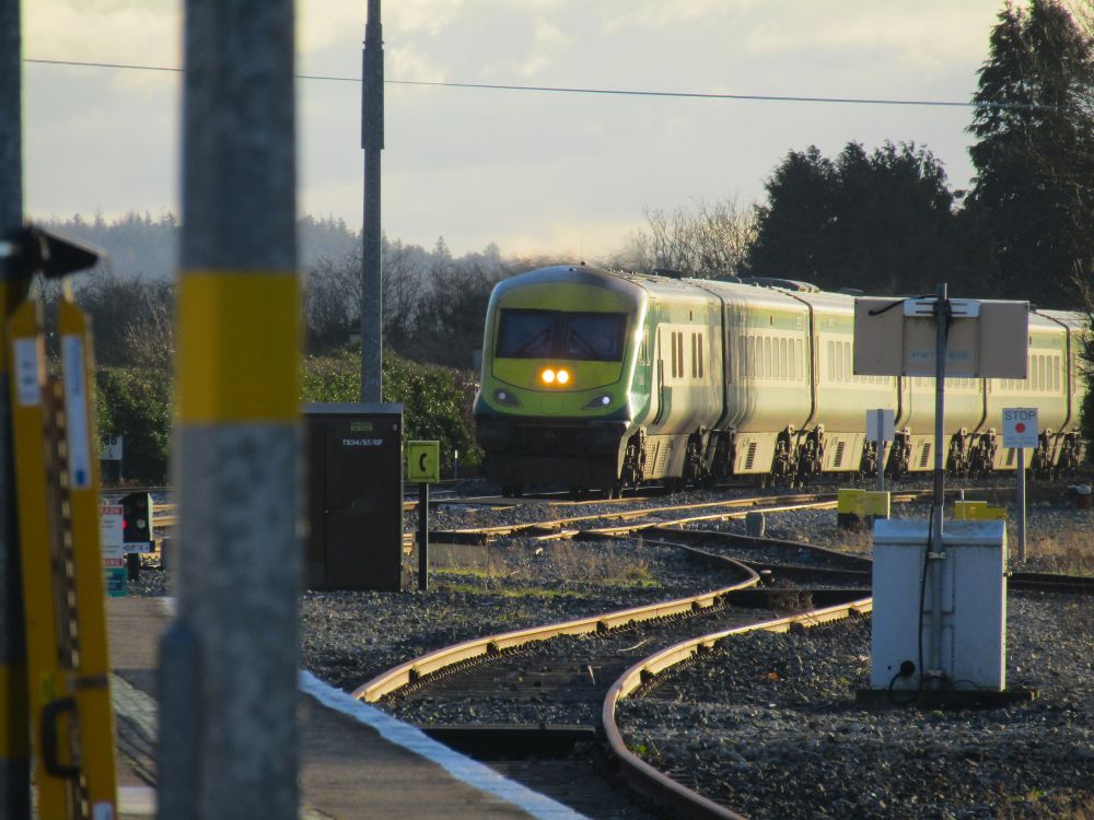 An Irish rail MKIV is approaching a station from a distance. It has its headlights and marker lights on. At the bottom of the photograph is some platform and rails
