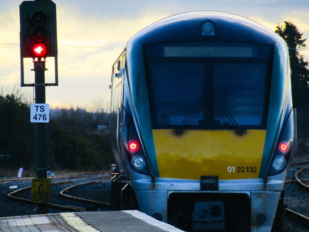 An Irish rail class 22000 DMU train has left a platform, in the foreground you can see the front of the train, some vegetation like hedges and a signal (TS466) which is showing a red danger aspect