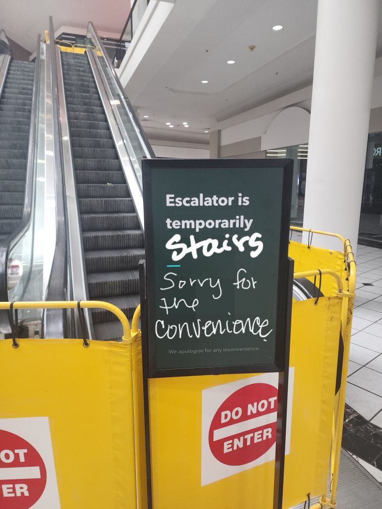 Picture of a sign in front of an escalator with the text "Escalator is temporarily stairs" with the text "Sorry for the convenience" under it.