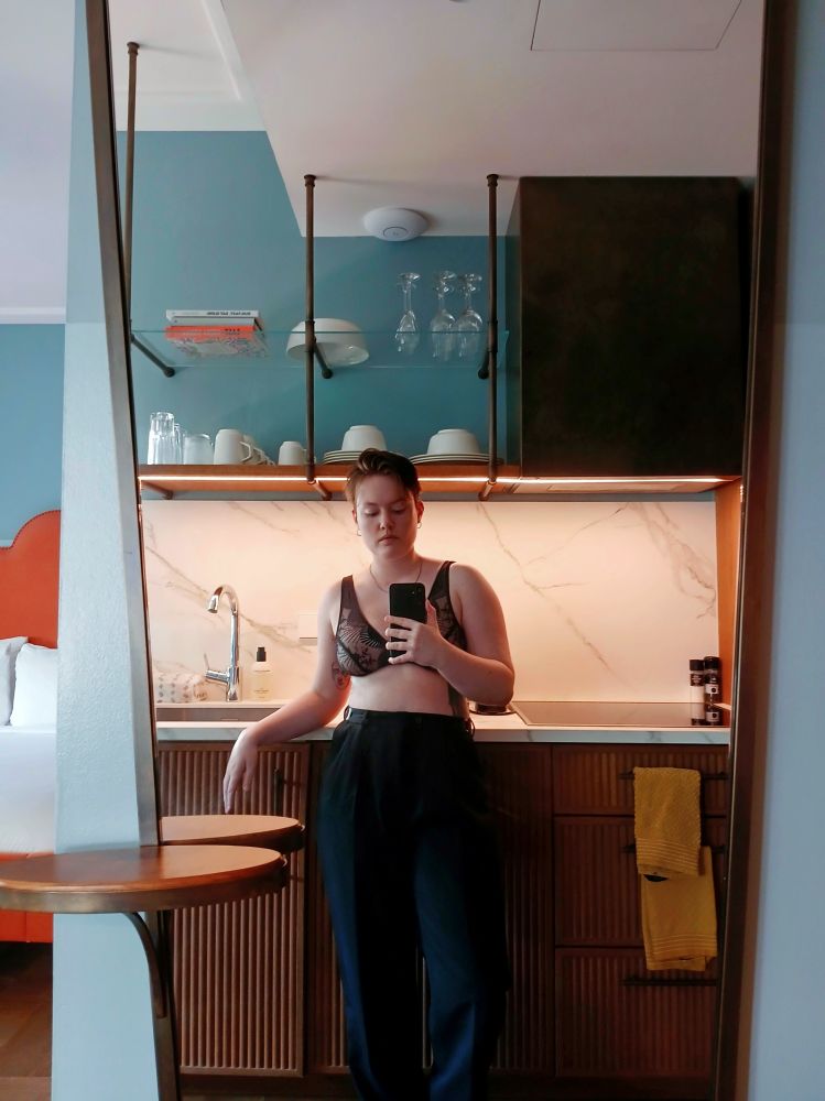 ash satine is taking a mirror selfie. She is wearing trousers and a bra, they have a tattoo on their right side. They're standing in a kitchen and the bed is reflecting in the mirror. 
