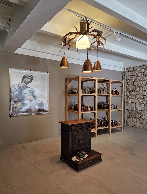 Blick in die Ausstellung
Im Vordergrund ist eine historische Gebetsbank aus Holz platziert. Darüber hängt eine moderne Pendelleuchte aus einem Hirschgeweih. Im Hintergrund ist ein Poster einer Schwarzweißfotografie von Michelangelos Medici Madonna zu sehen, an der Wand daneben steht ein Vorratsregal mit gefüllten Einmachgläsern. © Sandra Knecht, Foto: Sabrina Vogelbacher
