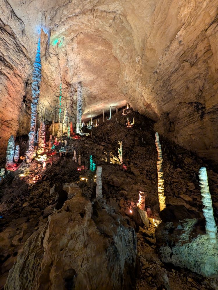 A closer shot deeper in the cave, looking up at towering stalagmites and stalactites, each illuminated in a rainbow of lights. The rough rock walls arch high overhead, emphasizing the sheer immensity of this BIIIIIIIG CAVE SYSTEM. 