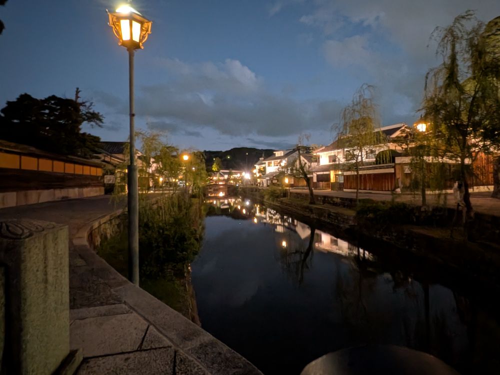A tranquil evening scene along the Kurashiki Bikan canal. Old-fashioned lamps glow softly, their light reflected in the still water bordered by willow trees and white-walled merchant houses. The sky is a deep blue fading into night, clouds drifting above the quiet historical street — serene, timeless, and gently illuminated.
