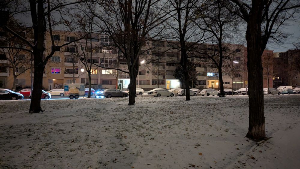 Eine Gründliche ist von frischem Schnee bedeckt. Parkende Autos sind ebenso schneebedeckt. Straßenlaternen beleuchten die Szene, während kahle Bäume und mehrstöckige Häuser die Straße säumen.