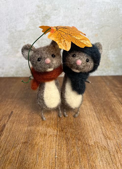 2 kleine mausbraune, weissbäuchige Herbstfilzmäuse mit Schal und eine davon mit Kappe unter einem Blatt-Regenschirm.
Gesehen bei Laura Statham (https://www.laurastatham.com)