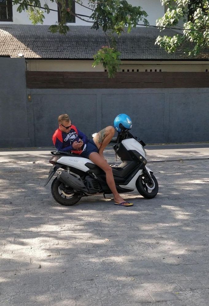 Foto de um cara reclinado numa moto, mas causando uma confusão visual como se tivesse um mulher de capacete sentada na moto com ele.