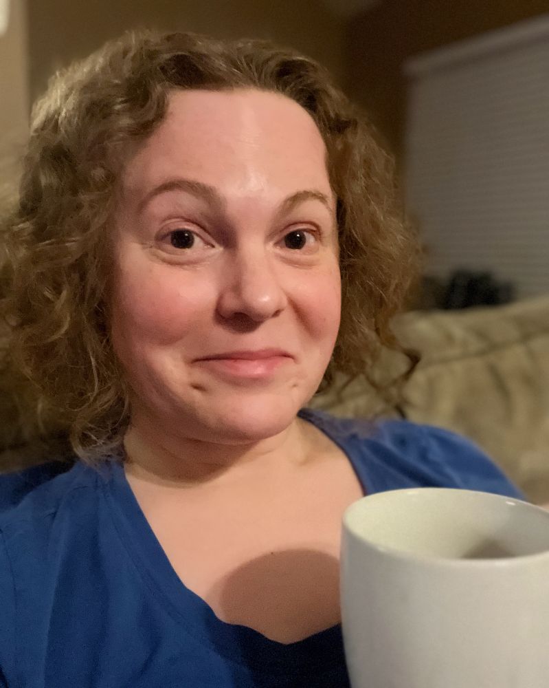 Middle aged woman with frizzy hair and a goofy look on her face, sipping coffee and wearing pajamas.