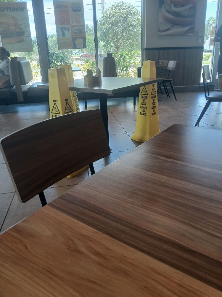 A photo taken within a Burger King restaurant where three yellow "caution, wet floor" signs are placed around a table in the center of the room. A Burger King crown sits on the table close to one of the signs. 