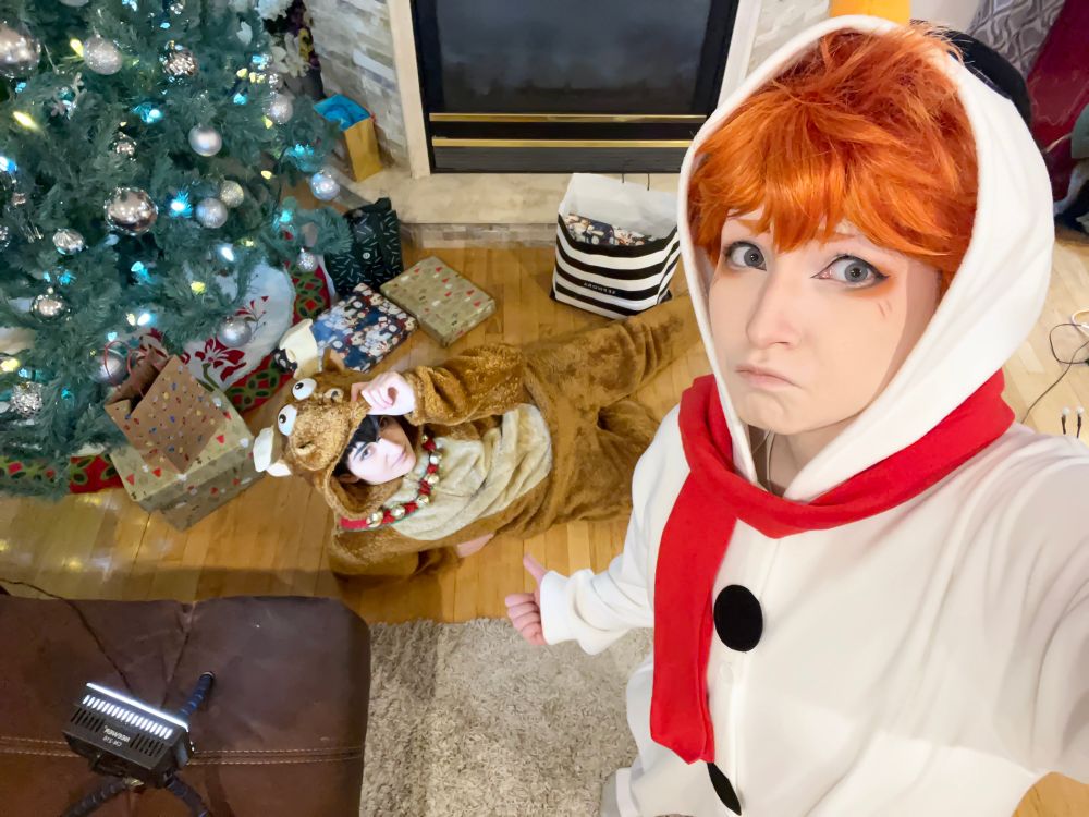 Tobio Kageyama and Shoyo Hinata, Haikyuu, in snowman and reindeer onesies. Kageyama is under a Christmas Tree as a present for Hinata for the holidays. This is cosplay.