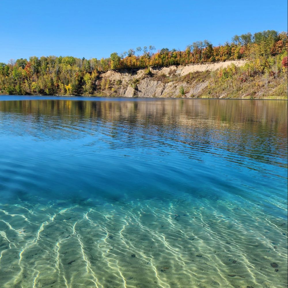 A sunny fall day in Northern Minnesota. This photo shows stunning tropical colored and clear blue water of a lake and its shore. The background shows a sandy, rocky cliff surrounded by fall-colored trees and a clear blue sky.