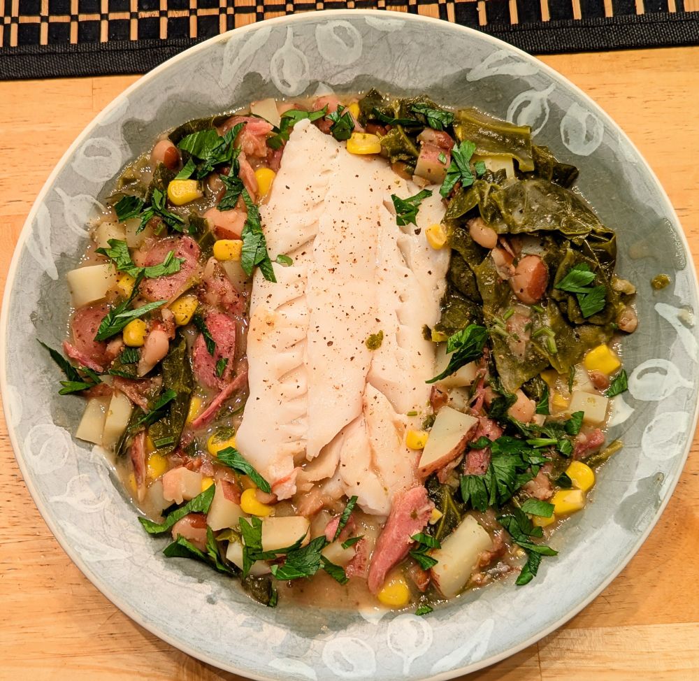 A light green bowl with a braise of beans, greens, corn, ham, potato, and cod. Garnished with parsley.
