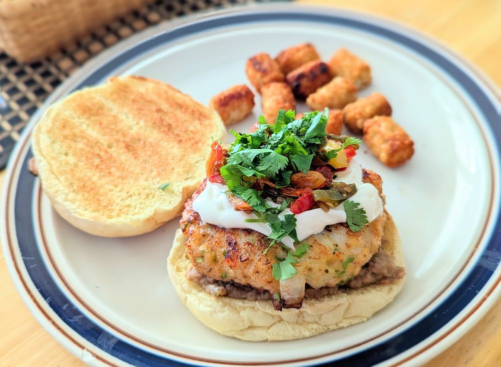 A white plate with a blue border holding a toasted burger bun topped with refried beans, a shrimp burger, mayo, roasted tomato salsa, and cilantro.  There is a small pile of tater tots in the background.