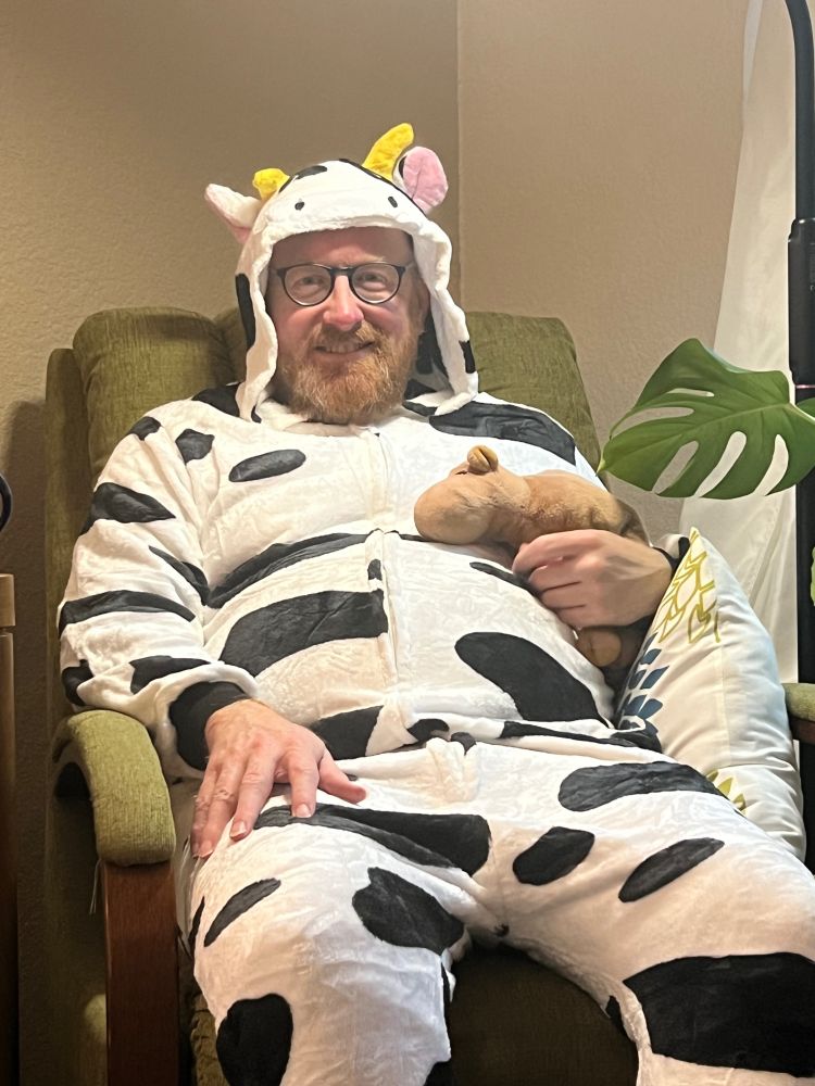 Matt sits in a chair, wearing a cattle onesie, and holding a stuffed Ty camel.