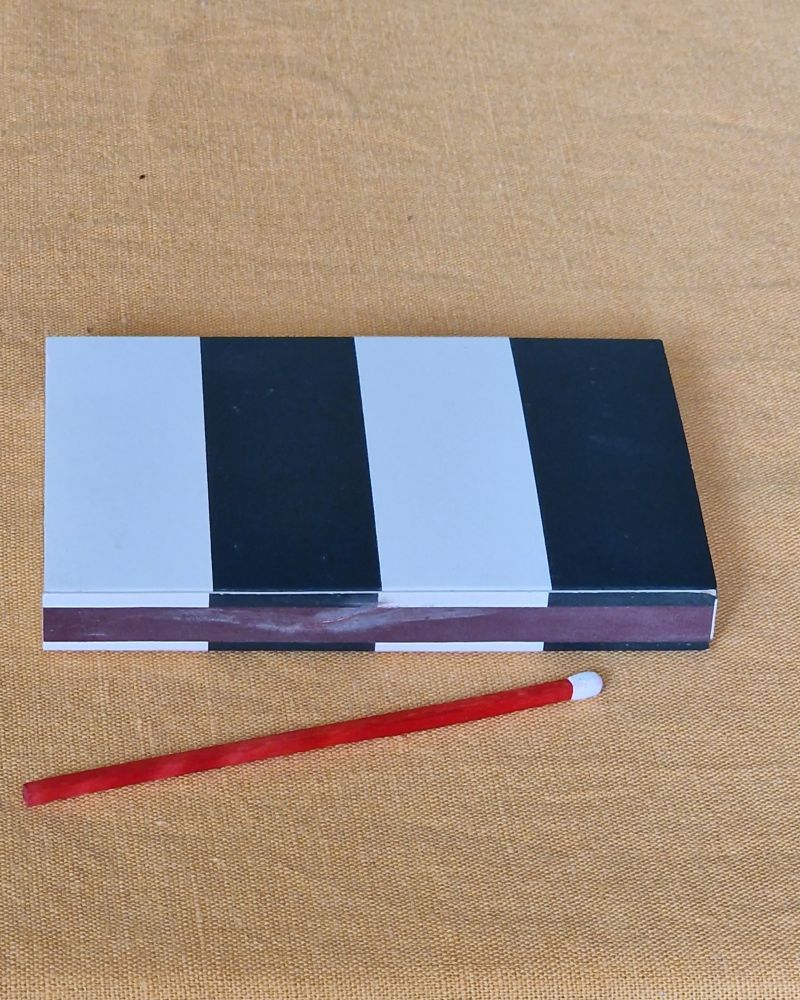 Scharz weiß gestreifte Streichholzschachtel, je zwei Streifen im Wechsel, liegt längs auf einer gelblichen Tischdecke. Davor liegt ein rotes längeres Streichholz mit weißem Kopf