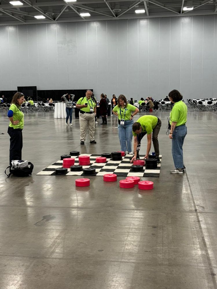 5 people in Green shirts gathered around a giant game of checkers.