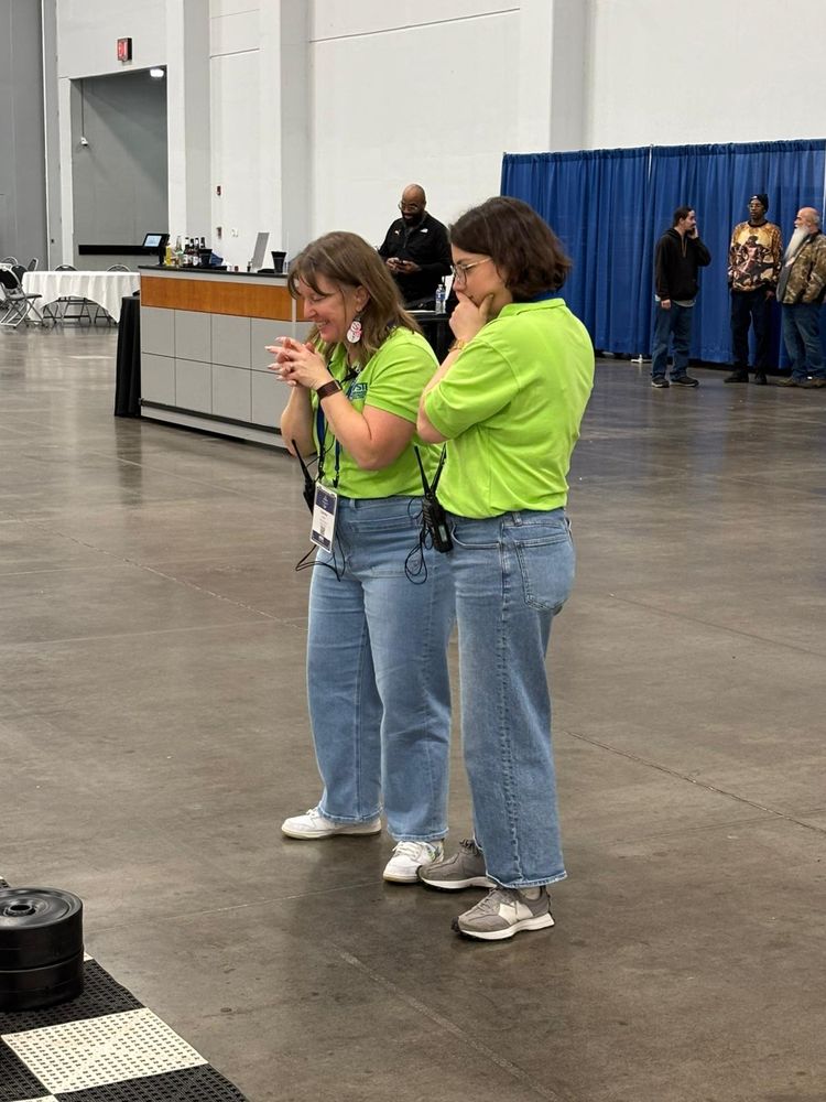 Two women wearing green shirts strategizing over an unseen giant checkers board.
