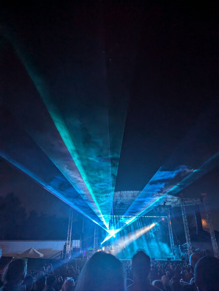 Blue lasers beaming out over the audience from a stage set against the night sky. A single white spotlight casts across the audience